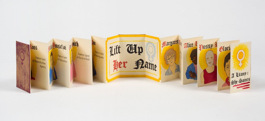 Photo of accordion book stretched open standing on table - center 3 pages read "Lift Up Her Name" in calligraphy - pages to left and right show ink illustrations of faces with names in calligraphy above