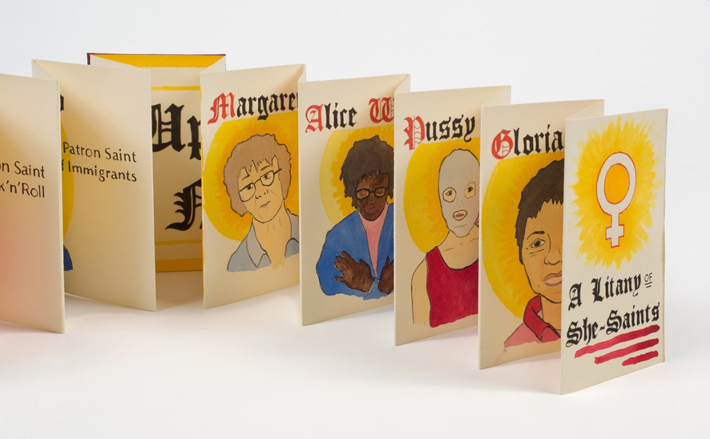 Photo of pages from accordion book standing open on table - partially visible illustrations and names: Margaret Crane, Alice Walker, Pussy Riot, Gloria Andzaldua